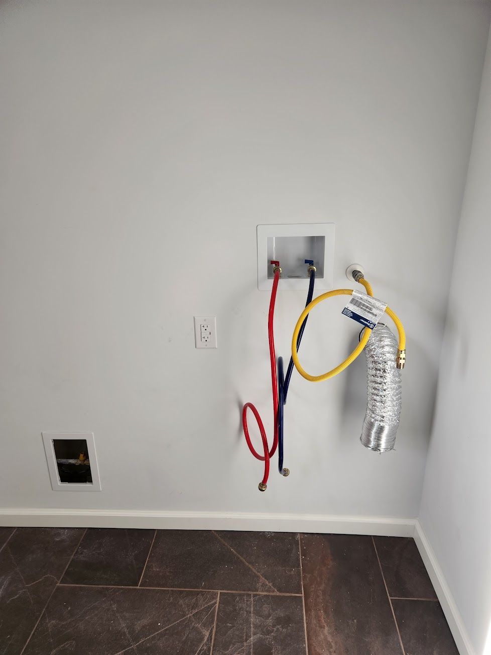 Laundry area wall with red and blue water hoses, a yellow gas line, a silver dryer vent, and electrical outlets.