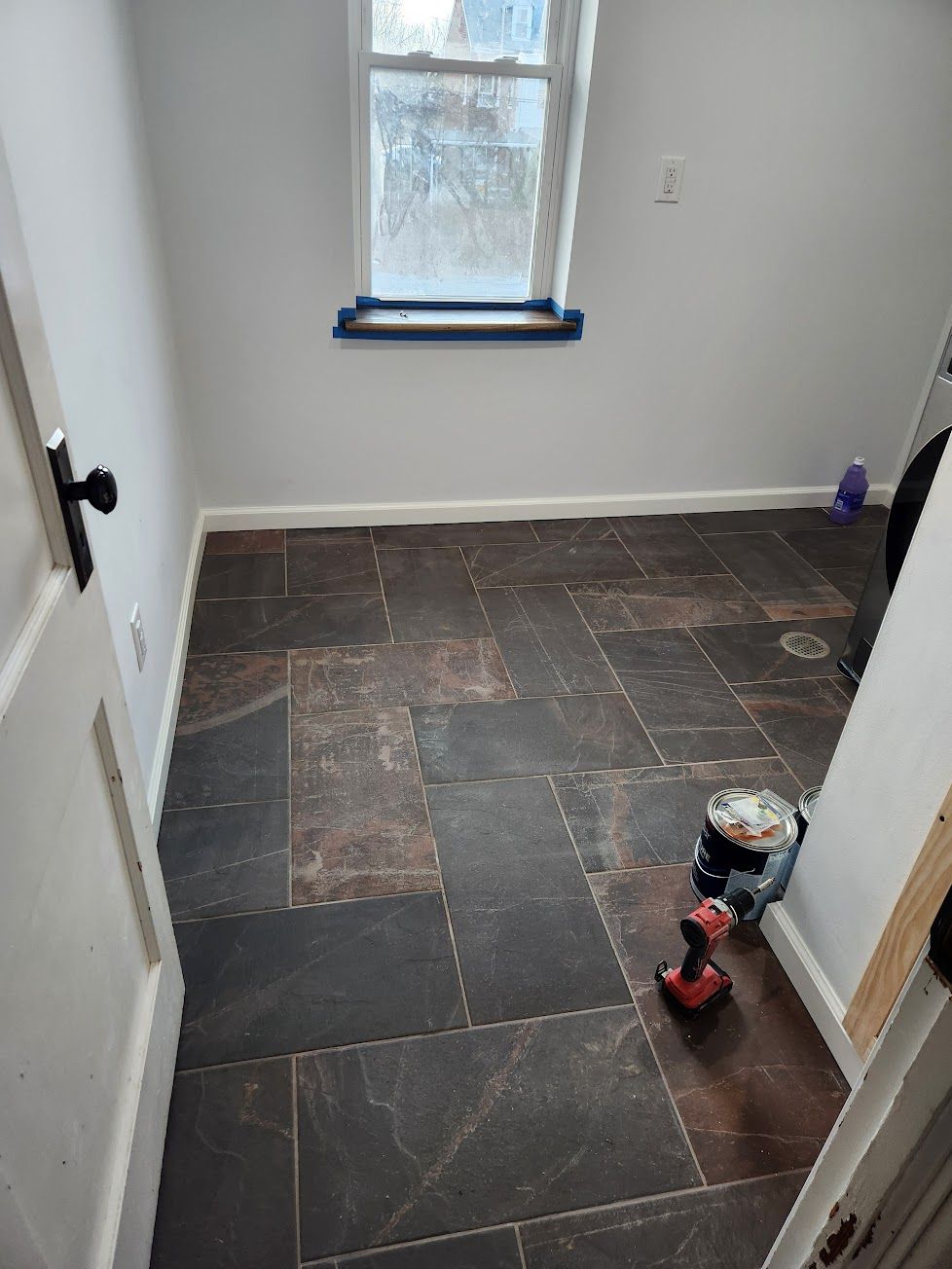 A room with new gray slate-style rectangular floor tiles laid in a herringbone pattern and a power drill on the floor.