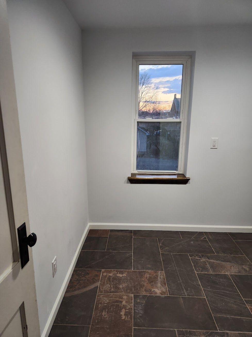 A small, empty room with light gray walls, a window overlooking a sunset, and dark, rectangular stone floor tiles.