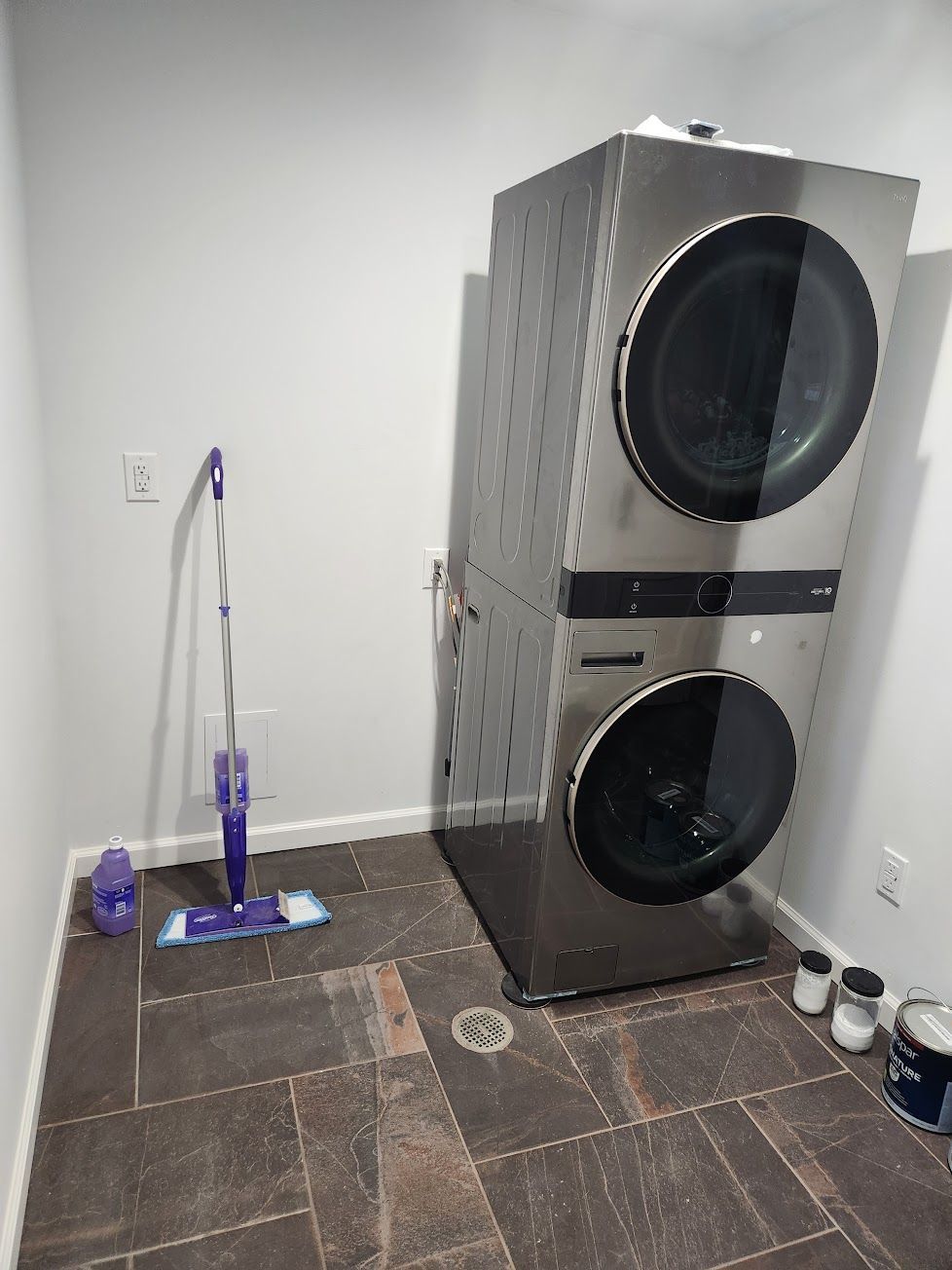 A laundry room with a stainless steel stacked washer and dryer unit, a purple mop, and floor tiles.