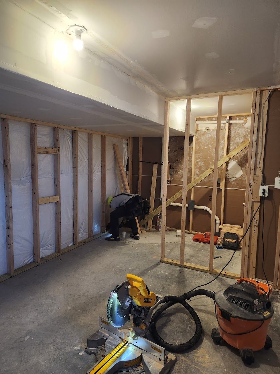 A construction worker builds a wood-framed wall in a basement, with a miter saw and shop vacuum in the foreground.