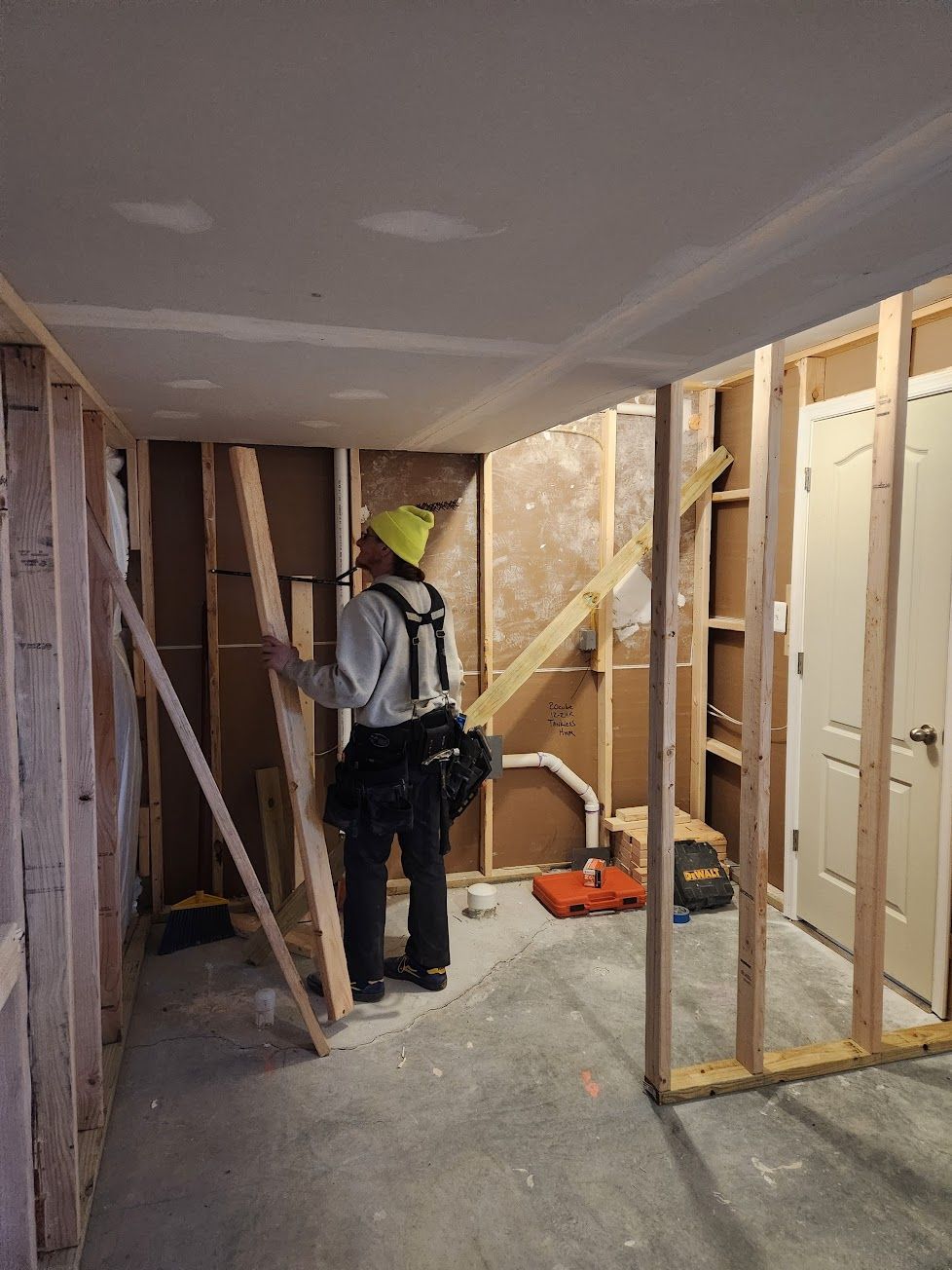 A person in a yellow beanie and tool belt works on the wooden framing of a room under construction.