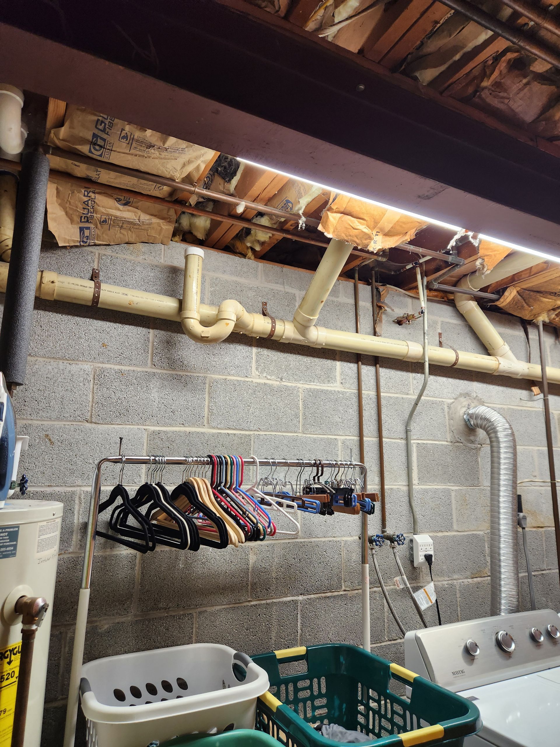 A basement laundry area with clothes on a rack, laundry baskets, and exposed plumbing pipes along a concrete block wall.