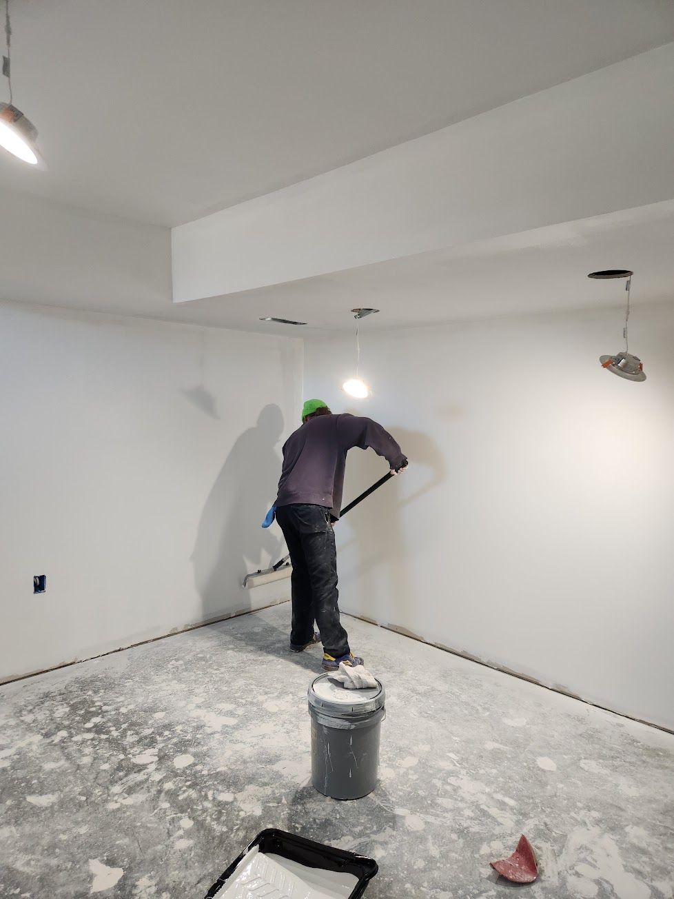 A person in a dark jacket and green cap uses a pole sander to smooth a white wall in a room under construction.