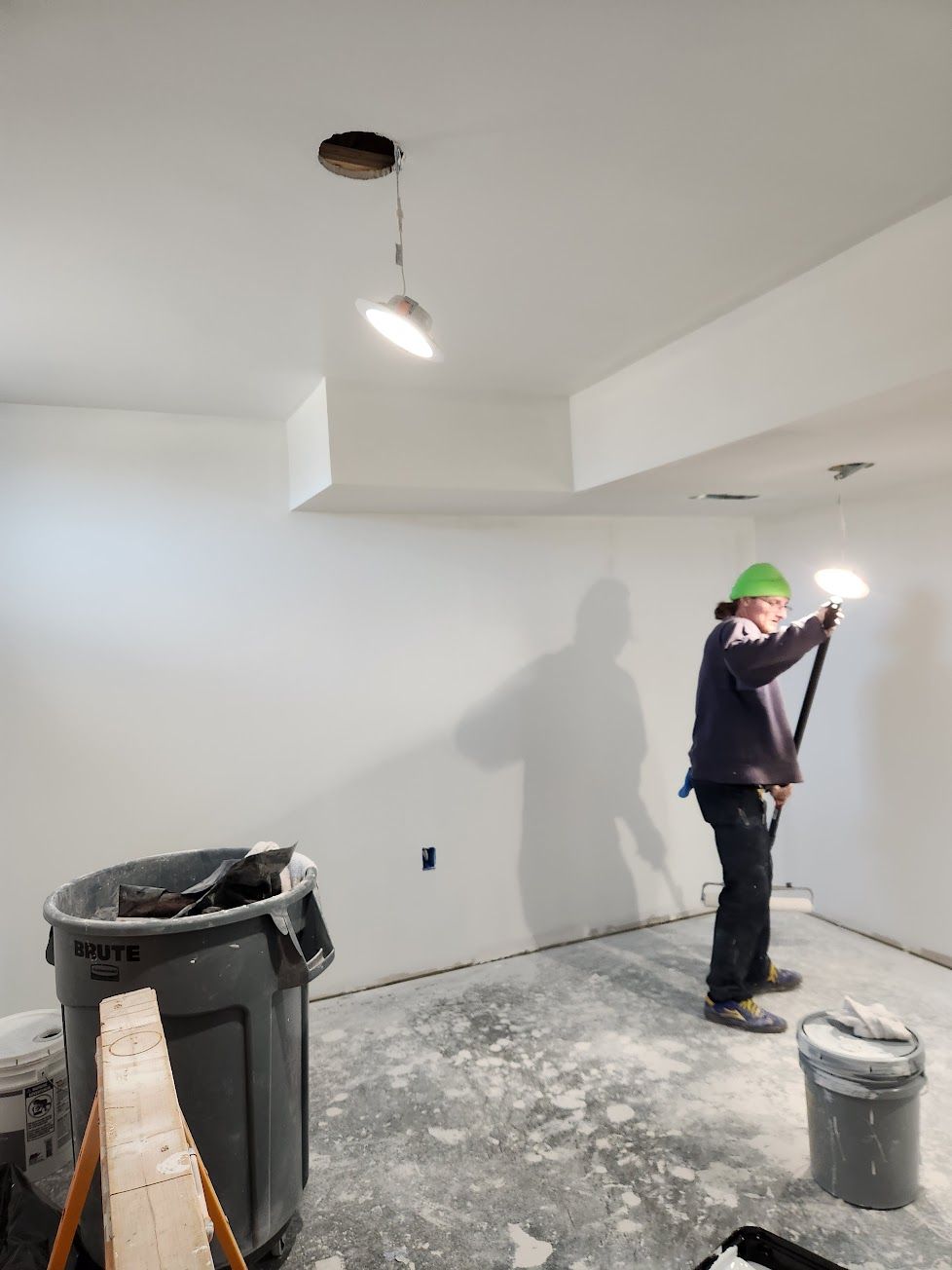 A worker in a green hat uses a long-handled sander on a white wall in a room under construction with two ceiling lights.
