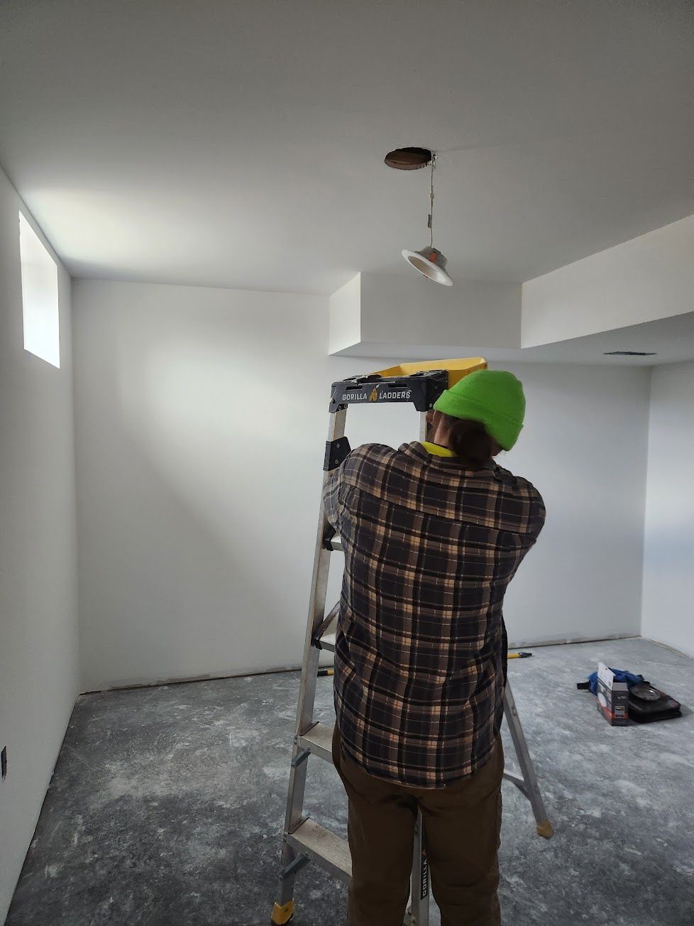 A person wearing a plaid shirt and green beanie stands on a step ladder, working on a ceiling light in an unfinished room.