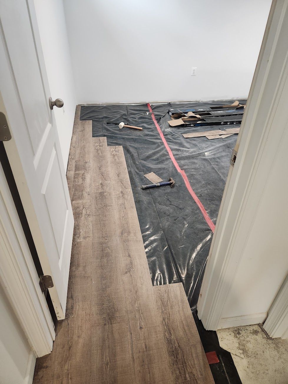 A view from a doorway of a room in progress with light wood-look planks installed over a black plastic underlayment.
