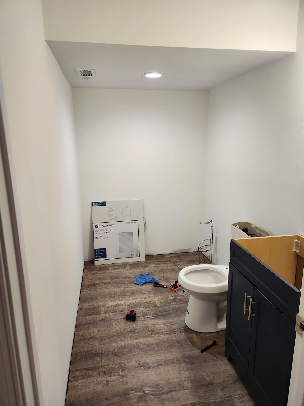 A bathroom under renovation with wood-look flooring, a toilet, a navy vanity cabinet, and an unmounted mirror in the back.