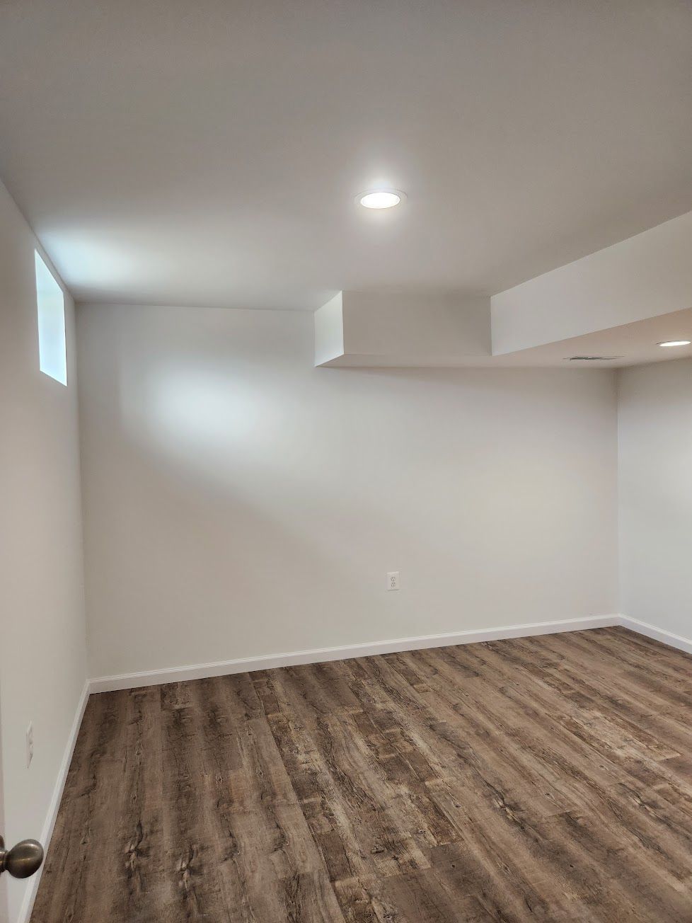 An empty room with white walls, a single recessed ceiling light, and brown wood-look plank flooring.