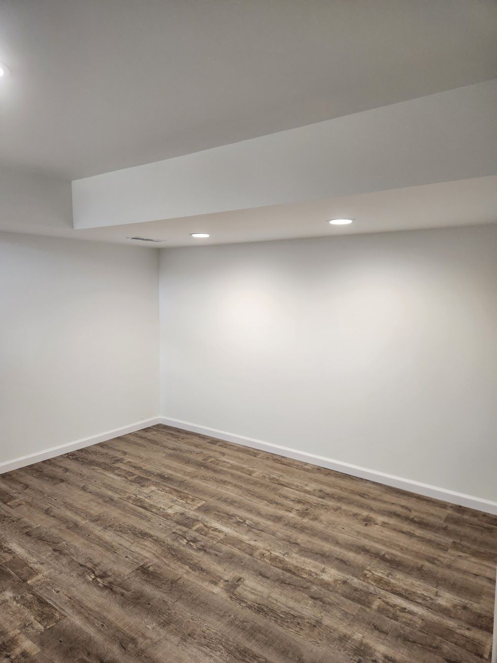 An empty room with white walls, light-colored wood-look flooring, and a recessed ceiling with two small lights.