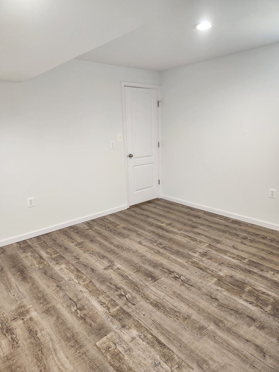 A bare room with white walls, light wood-look laminate flooring, and a single closed white door in the corner.