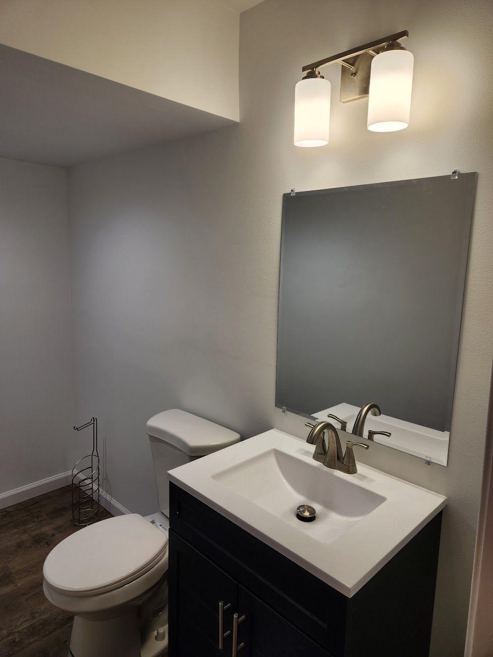 A modern half-bathroom with a white toilet, dark vanity with a white sink, rectangular mirror, and two-light fixture.