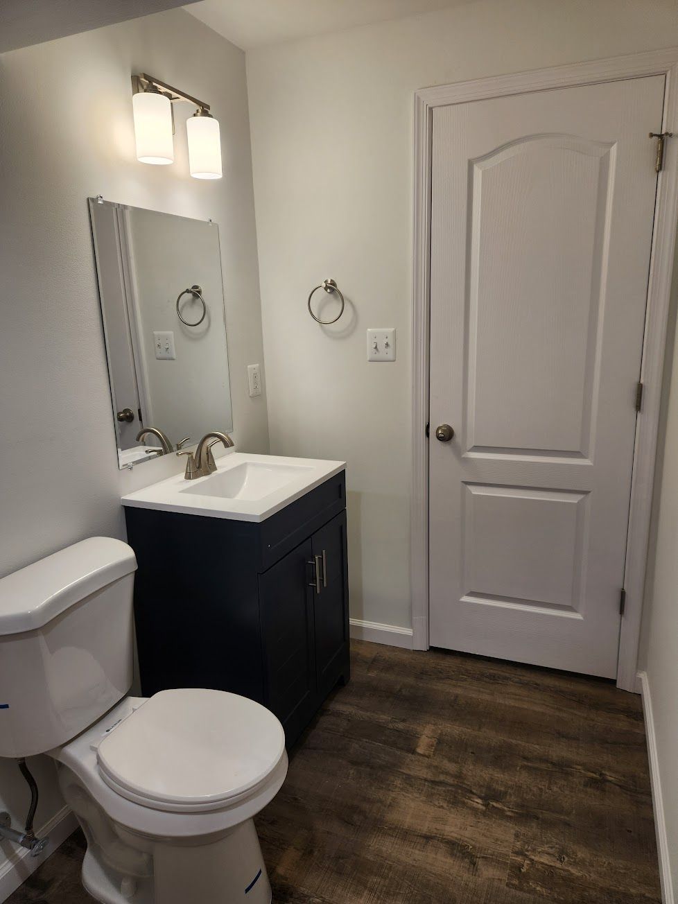 A small bathroom with a white toilet, a navy blue vanity with a white sink, a mirror, wall light, and a white interior door.