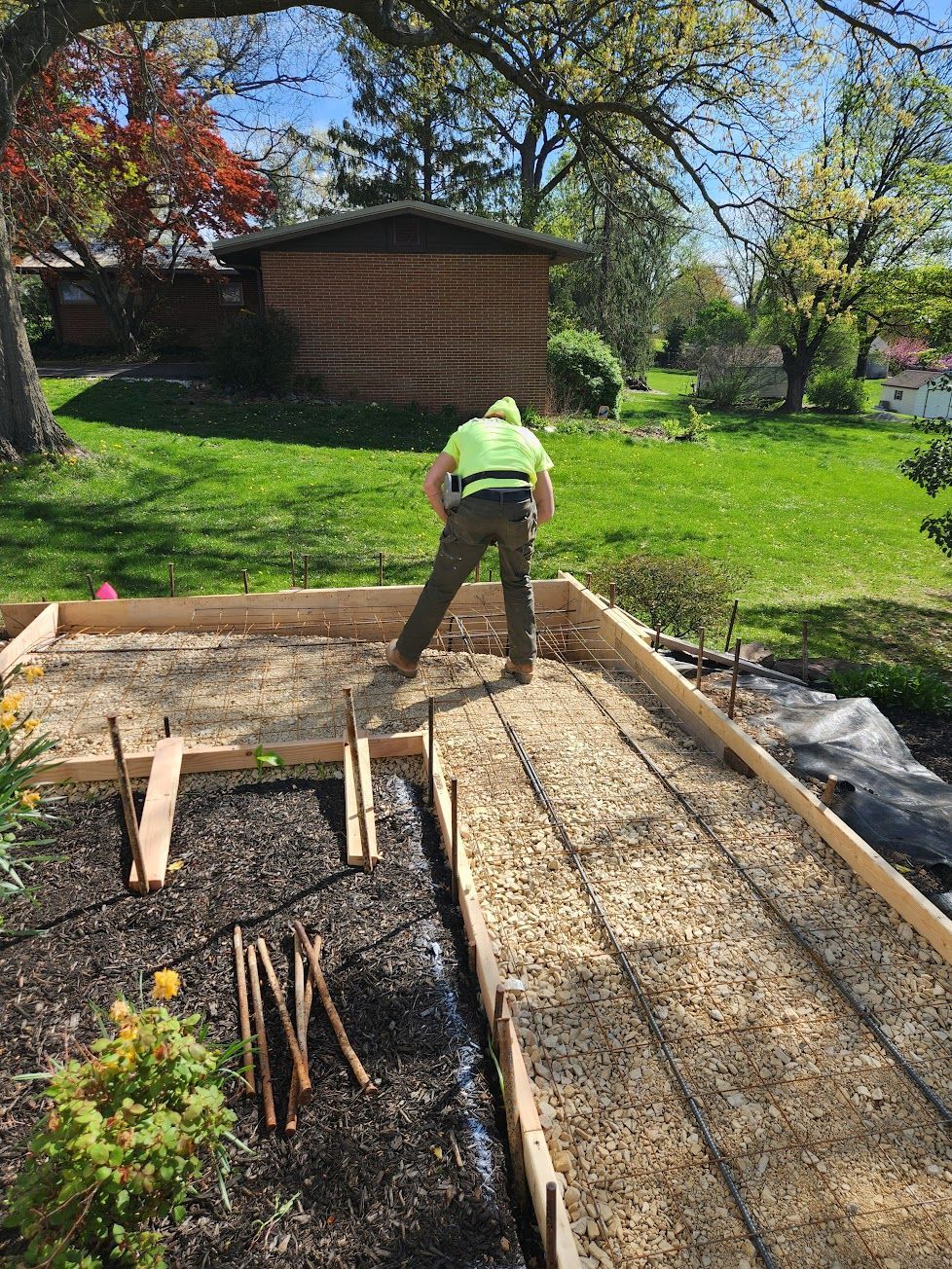 A person wearing a bright yellow safety vest works on a landscape project in a sunny backyard with wooden garden borders.