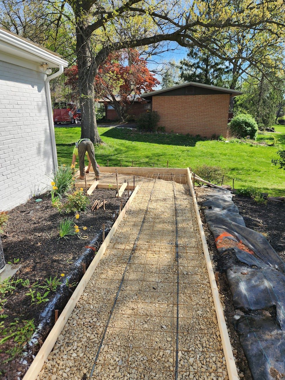 A person works on a wooden-framed, gravel-filled path for a new walkway next to a white brick house and a brick garage.