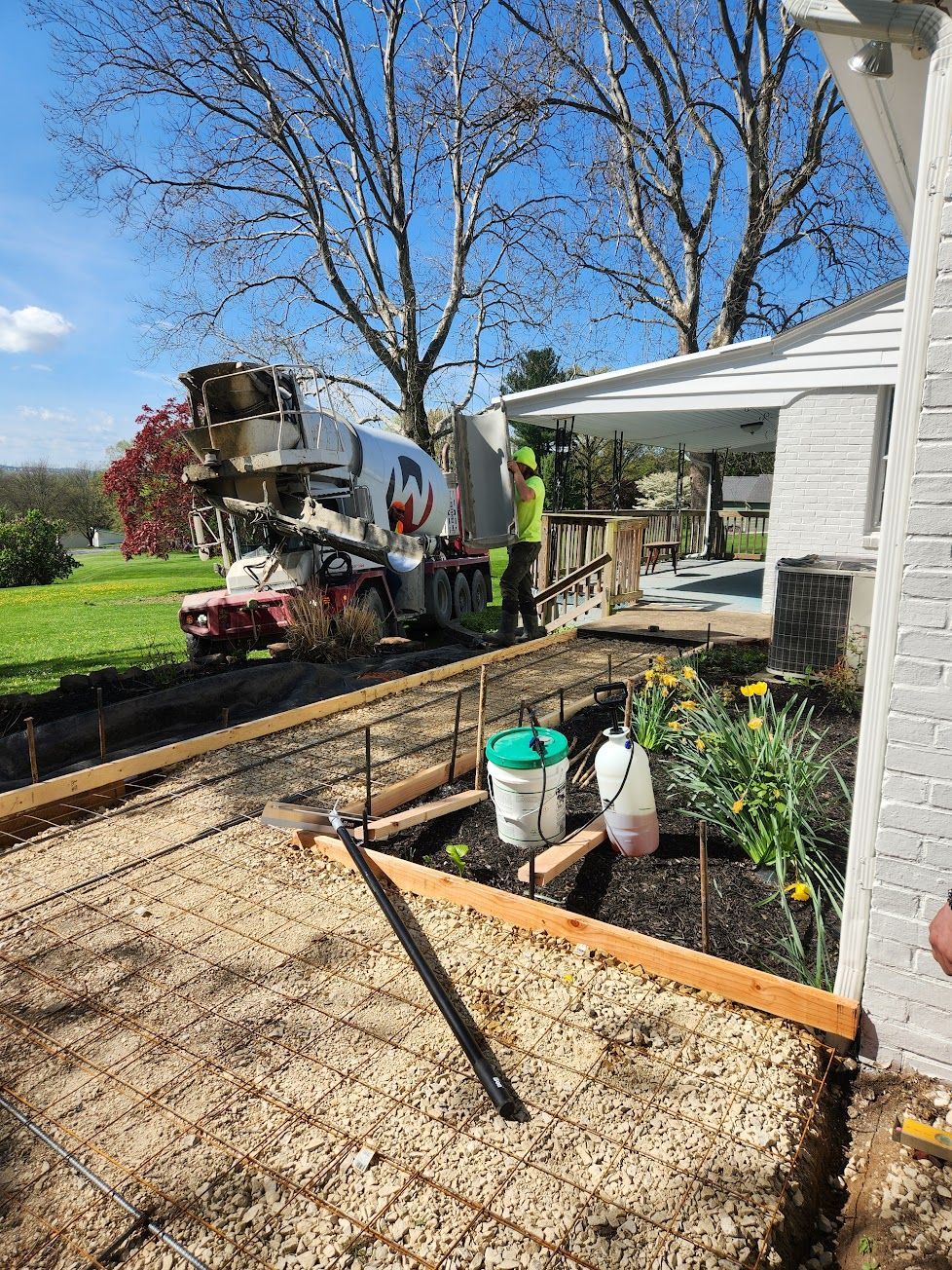 A cement truck pours concrete for a new walkway along a residential building next to a flower bed under a bright blue sky.