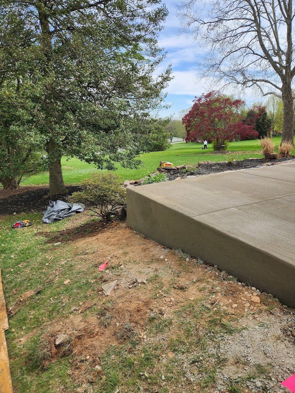 A concrete patio edge slopes down to a yard with trees, a red bush, and some construction debris in the grass.
