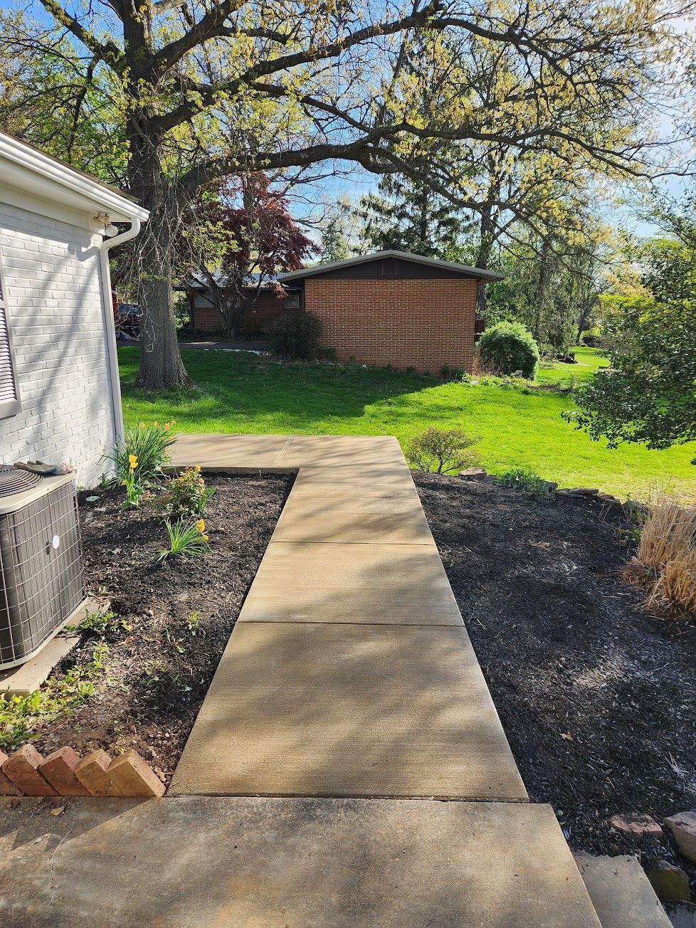 A concrete walkway leads through a landscaped yard toward a brick structure under large trees on a sunny day.