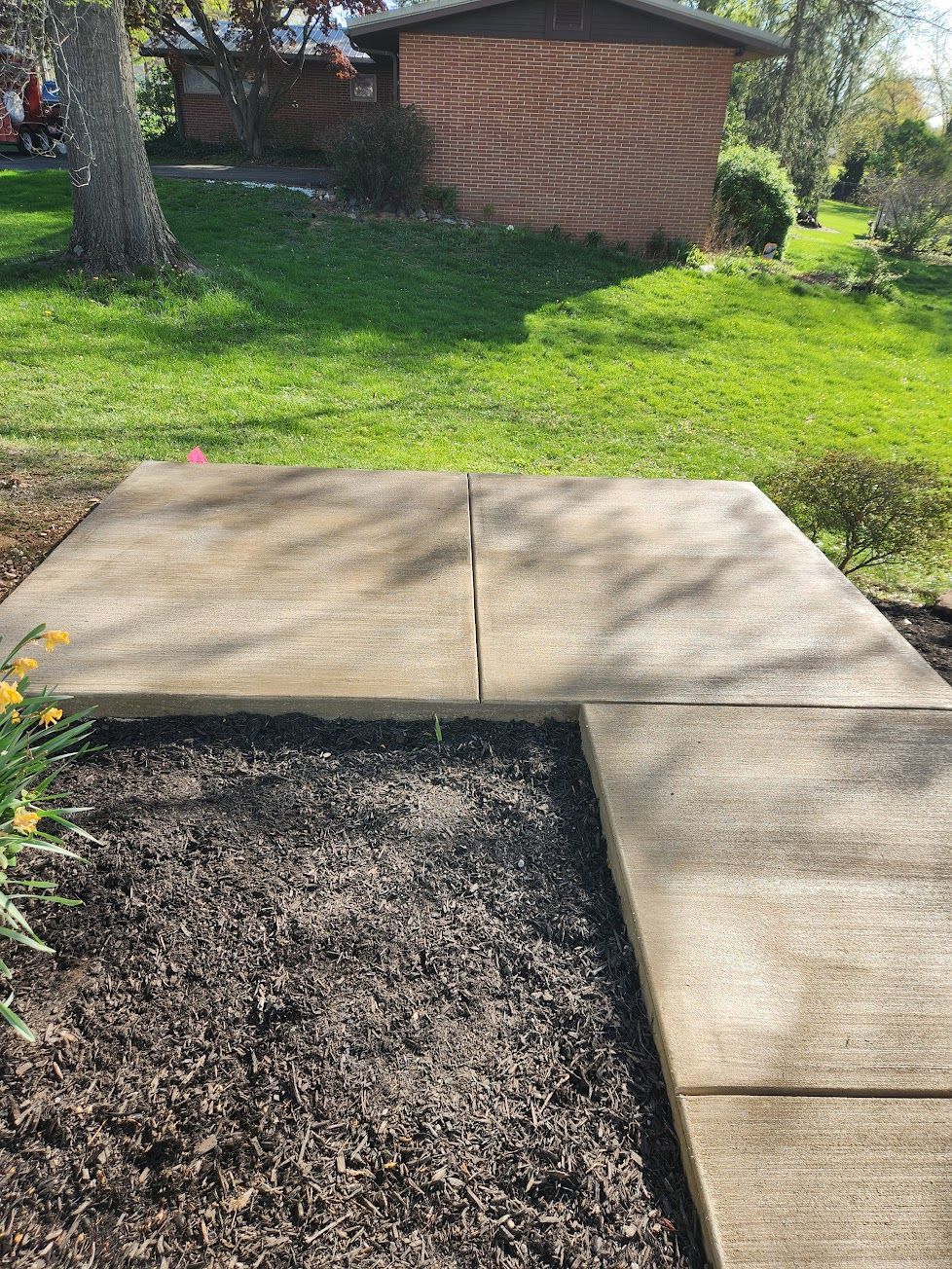 A concrete patio area set in a green backyard with dark mulch in the foreground and a brick house in the background.
