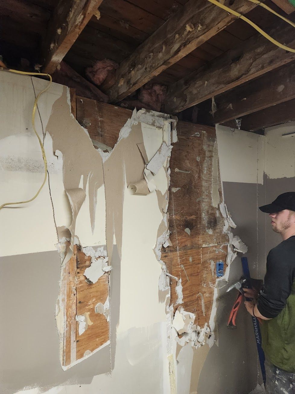 A person using a tool to remove damaged plaster and lath from a wall in a room with exposed ceiling joists.
