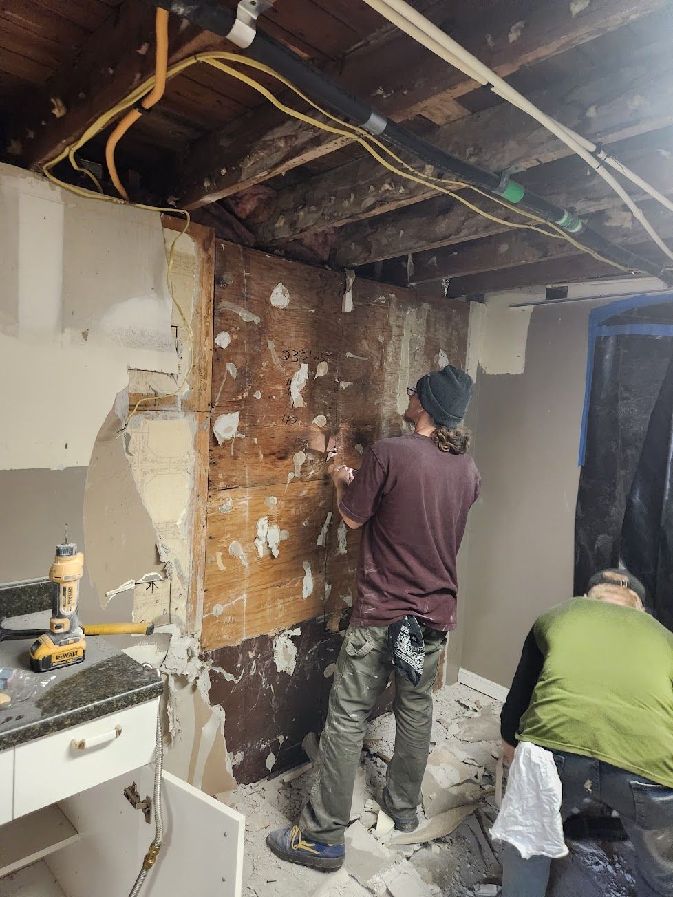 Two people work on a home renovation, stripping plaster from a wooden wall in a room with exposed ceiling joists.