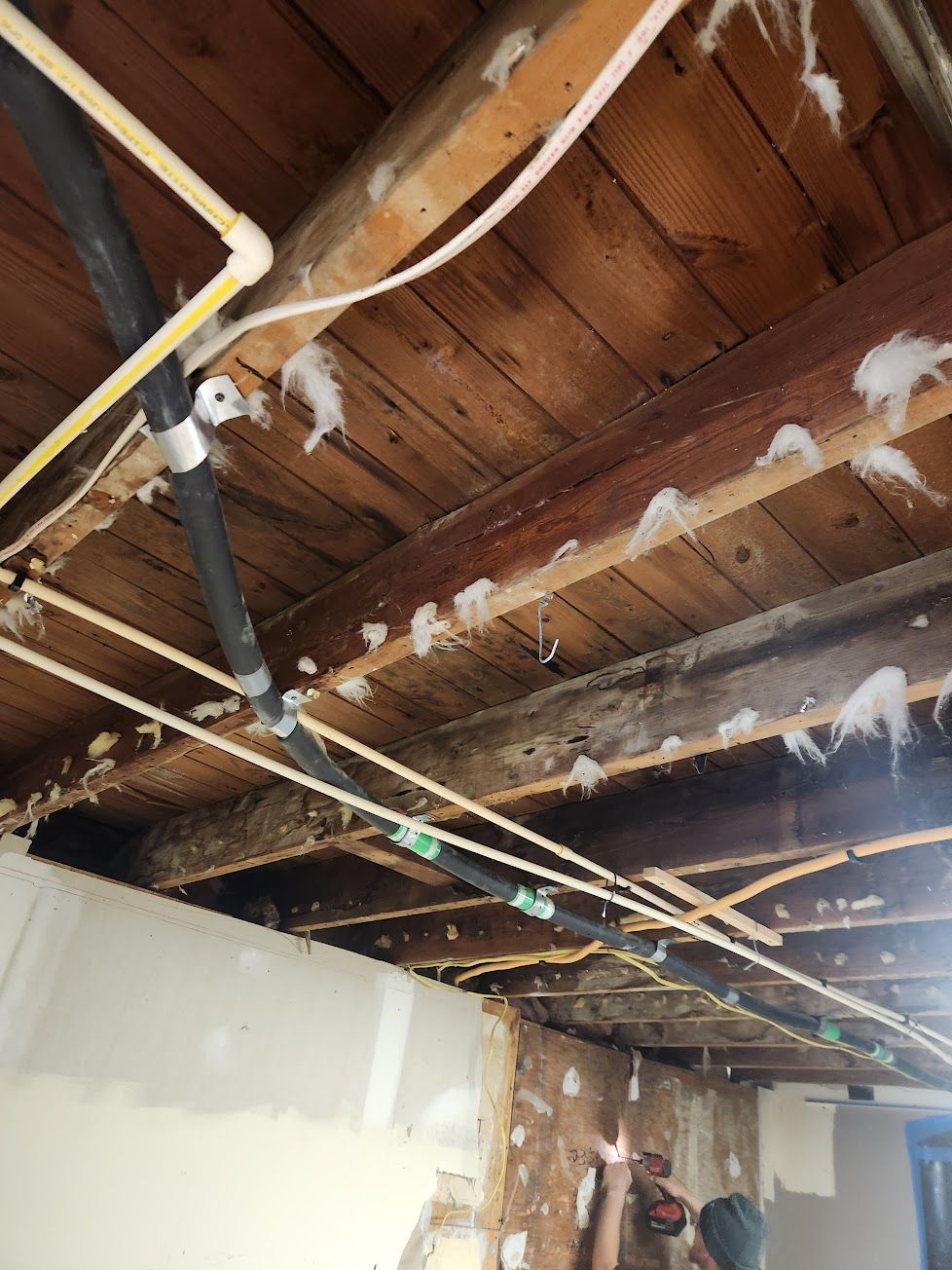 Exposed wooden ceiling joists with various pipes and wires running through them, showing construction in progress.