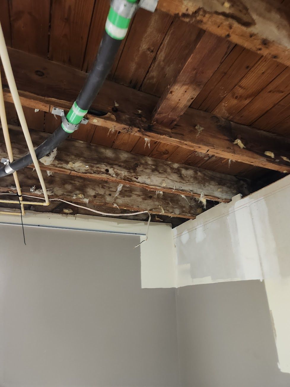 Exposed wooden floor joists and subflooring above a partially painted grey wall with a pipe running across the ceiling.