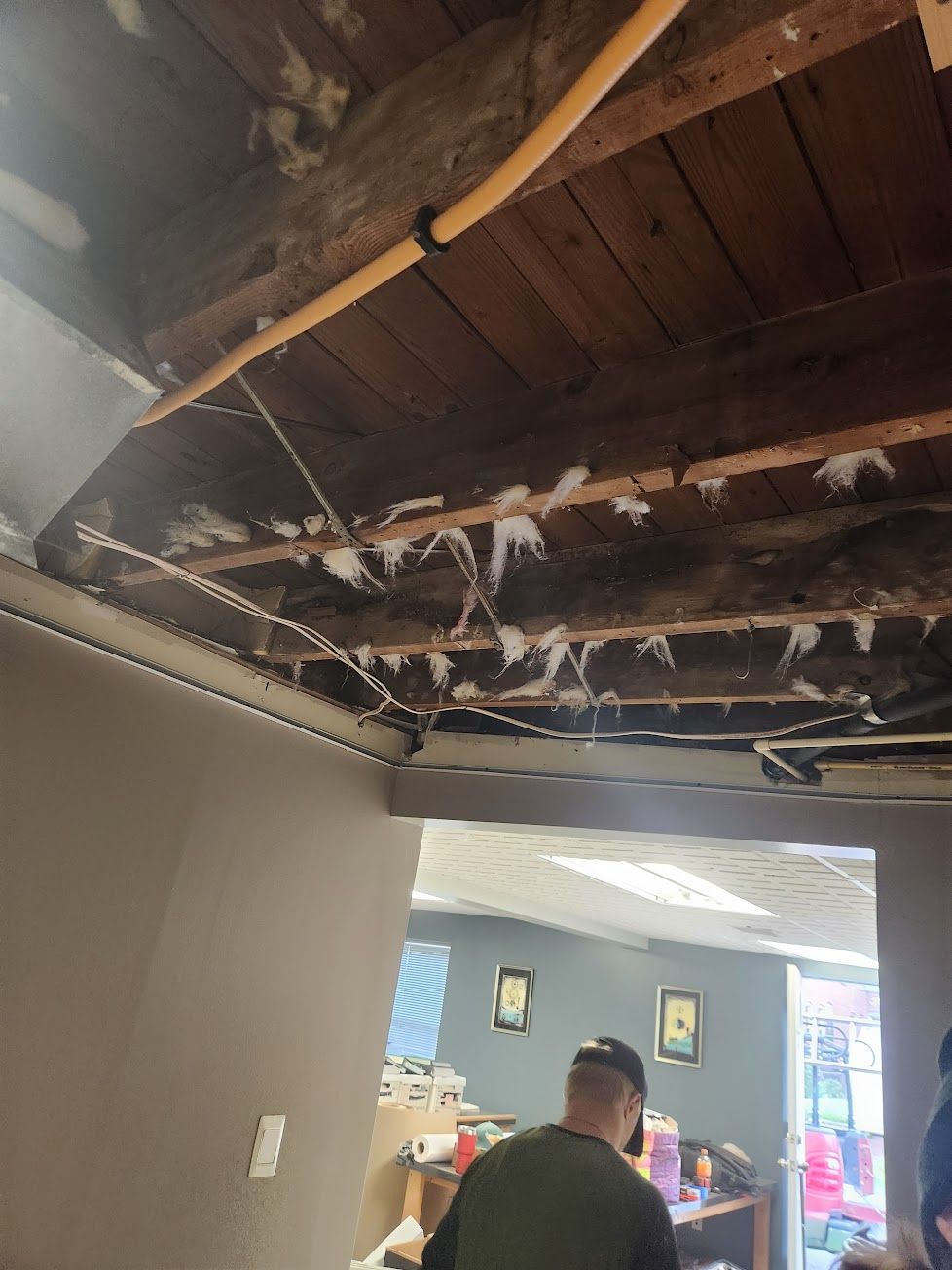 Exposed ceiling beams with hanging insulation and wiring, looking into a room where a person stands at a desk.