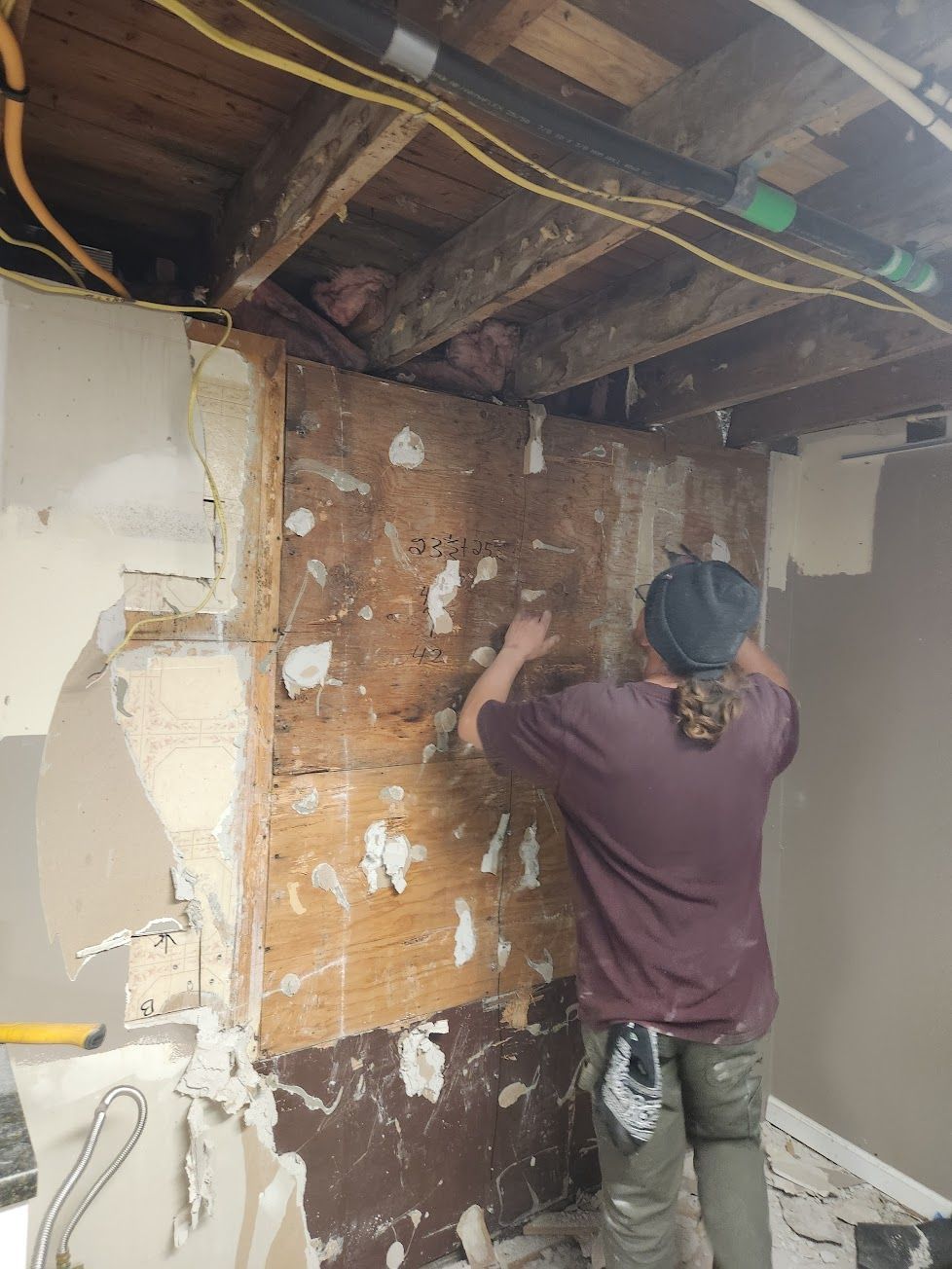 A person in a maroon shirt works on removing wall paneling in an unfinished room with exposed ceiling joists and wiring.