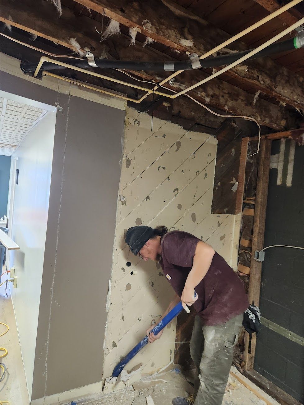A person in a maroon shirt uses a blue pry bar to remove plaster from a wall in a room under construction.