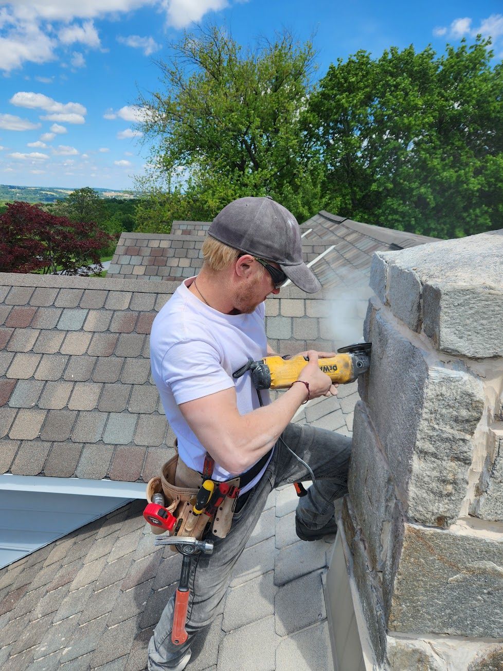 A worker wearing a cap and safety glasses uses a power tool to cut into a stone chimney on a residential roof.