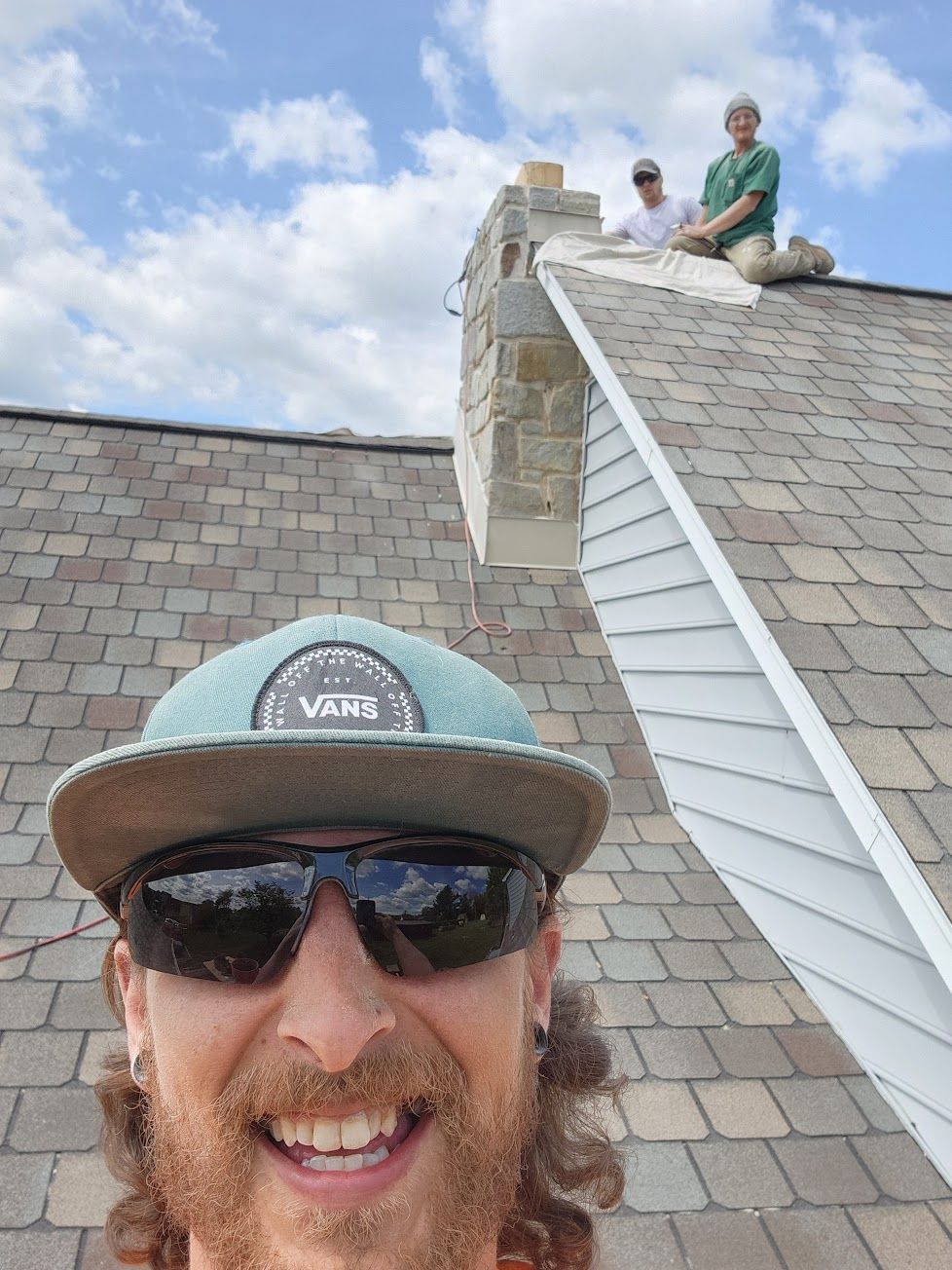 A person wearing a teal cap and sunglasses smiles for a selfie on a roof, with two other people working on the chimney.