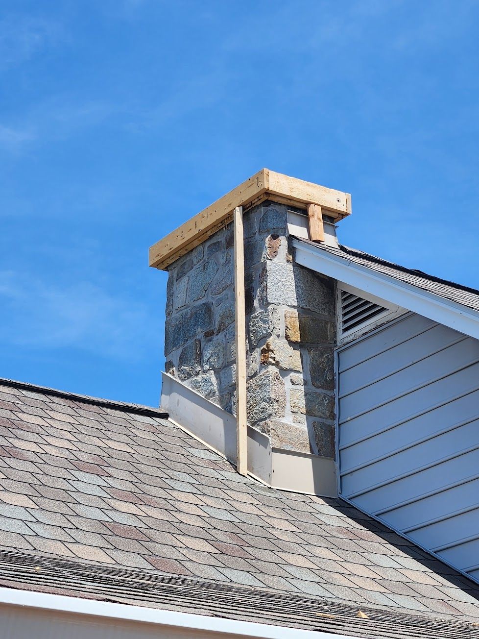 A stone chimney stack on a residential roof features a temporary wooden form installed at the top against a blue sky.