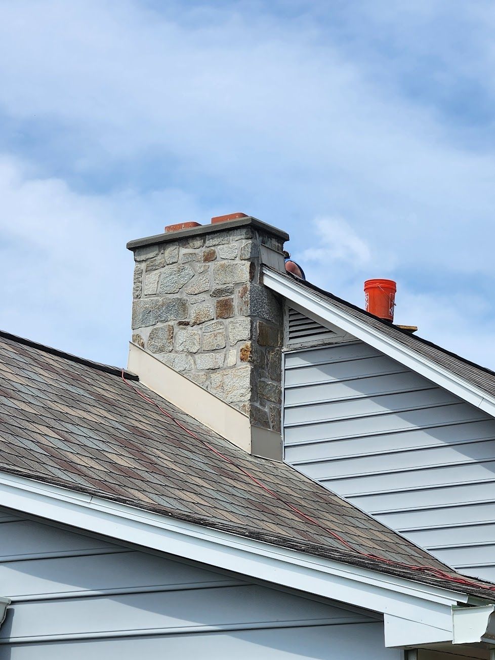 A stone chimney rises between two roof sections of a grey-sided house, with a small orange bucket placed on the roof.