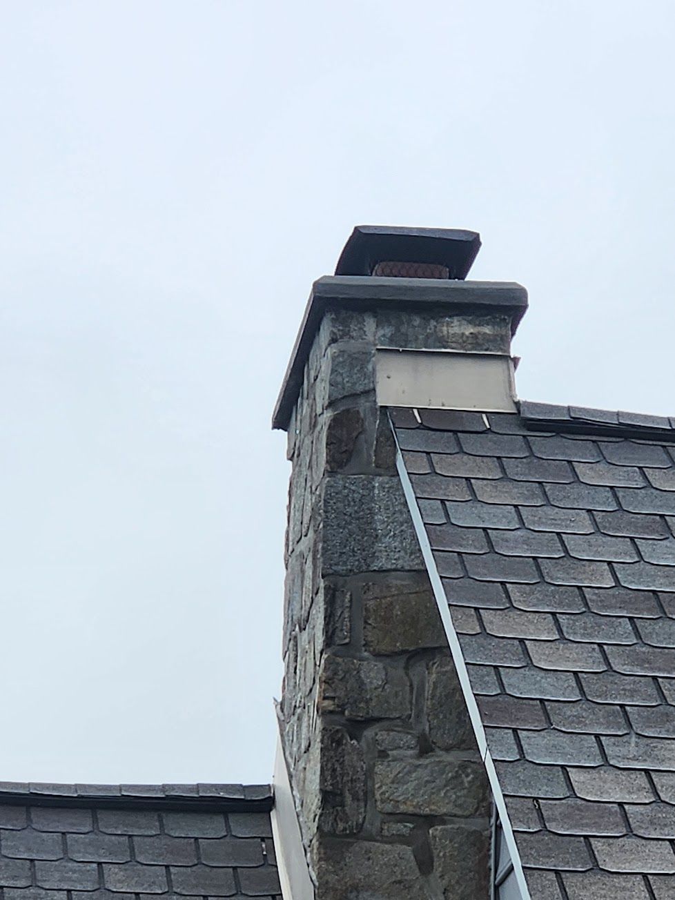A stone chimney stands against a pale sky, with gray roof shingles visible on the roof below.