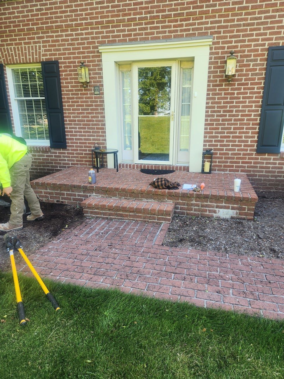 A worker in a neon jacket stands by a brick stoop and walkway leading to a glass storm door on a brick home.