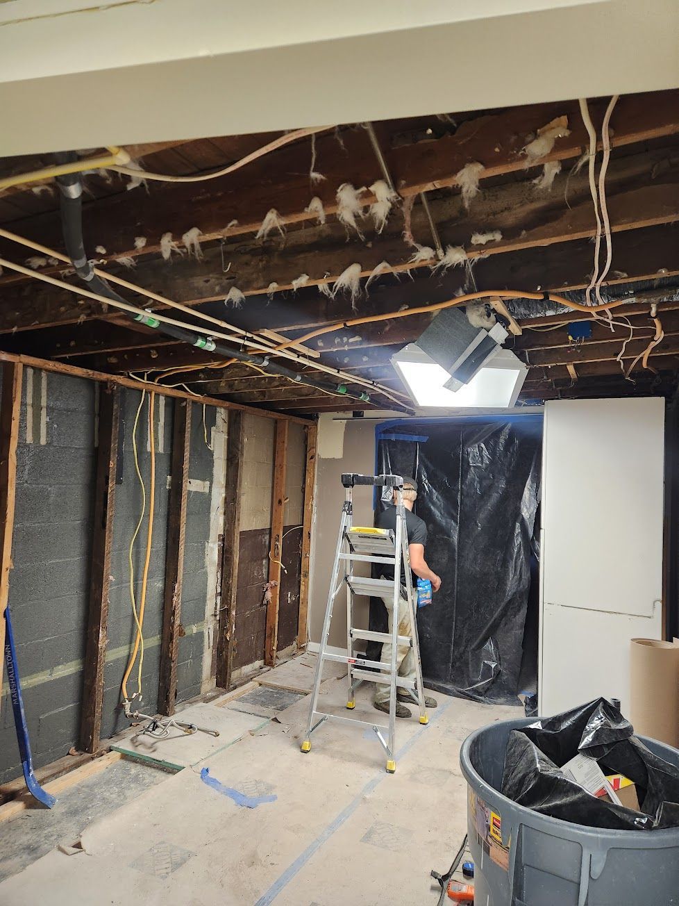 A person on a ladder in a room under construction with exposed ceiling beams, wall studs, and black plastic sheeting.