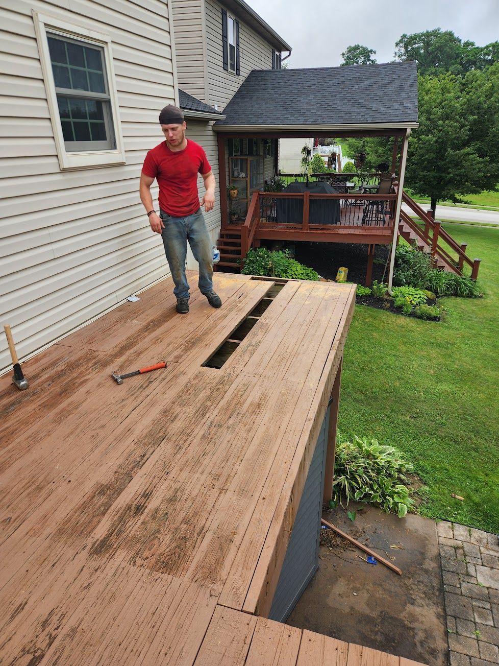 A person in a red shirt standing on a wooden deck with a gap in the floorboards under repair near a house.