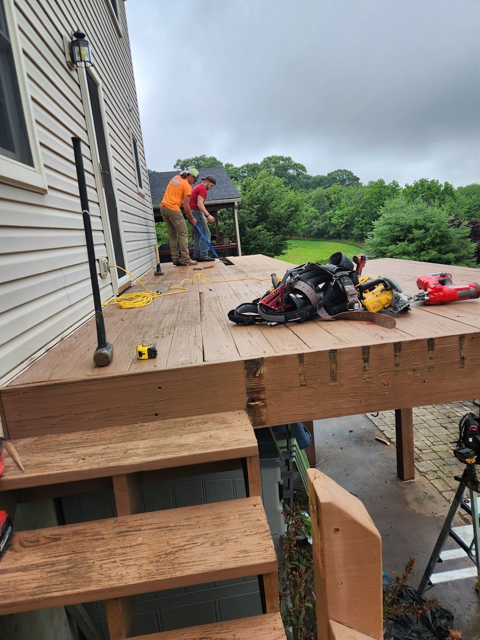Two workers build a wooden deck attached to the side of a house, with tool belts and gear resting on the surface.