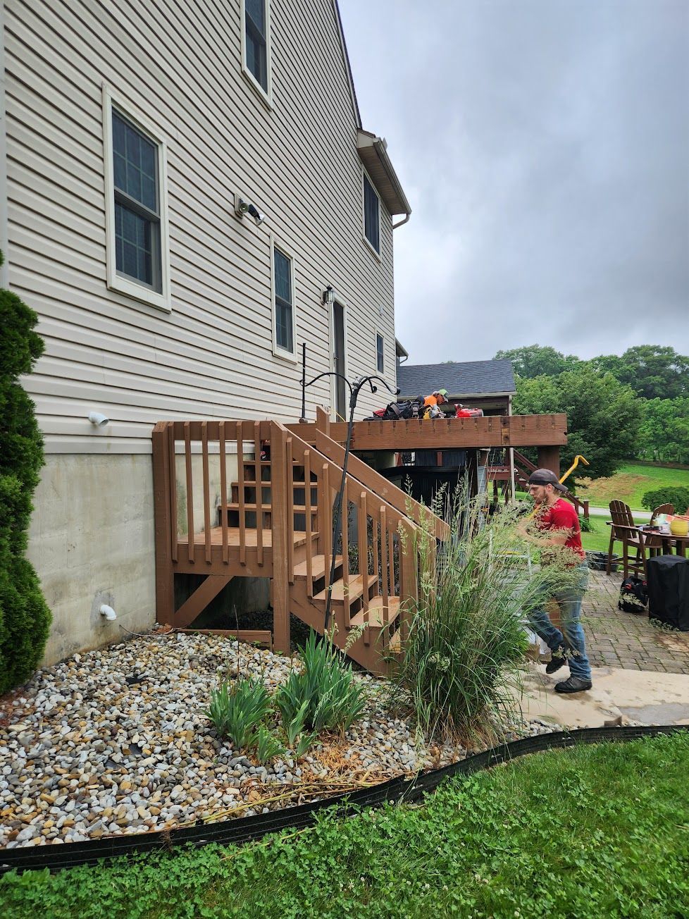 A person in a red shirt walks past wooden deck stairs attached to the side of a beige house with a gravel garden bed.