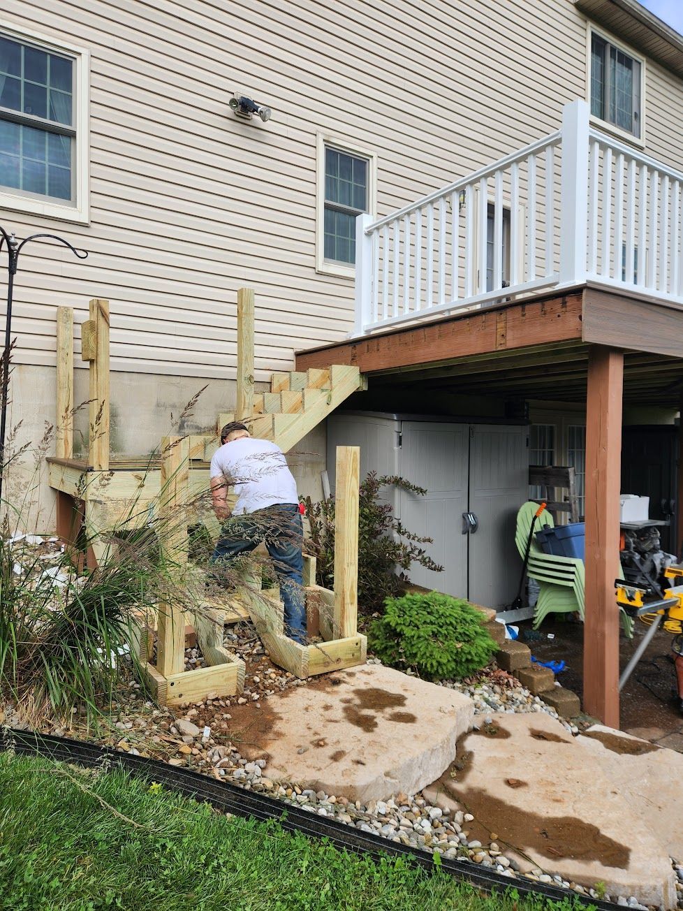 A person works on the wooden frame of a staircase being built against the side of a tan house with an elevated deck.