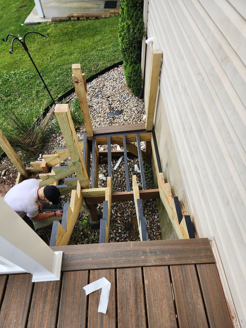 A worker builds wooden stairs leading down from an elevated deck onto a gravel-covered yard next to a house.