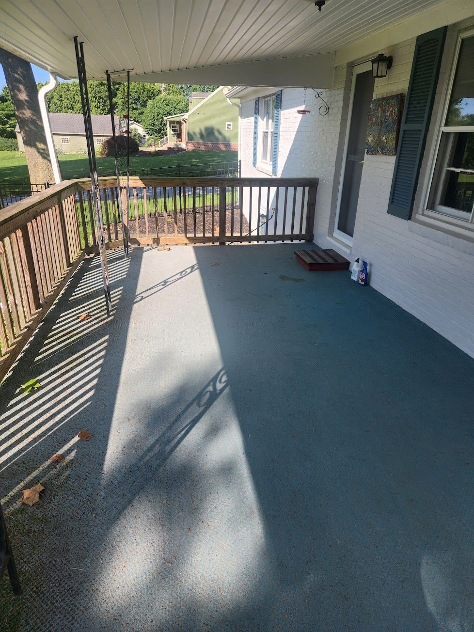 A gray-floored porch with a wooden railing, a hanging chair, and a white house exterior on a sunny day.
