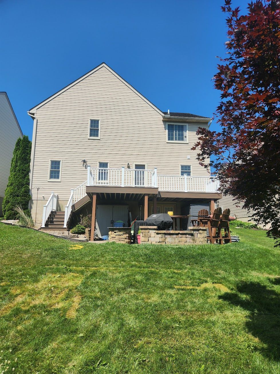 A two-story tan house with a raised wooden deck and stone patio area in a grassy backyard under a clear blue sky.