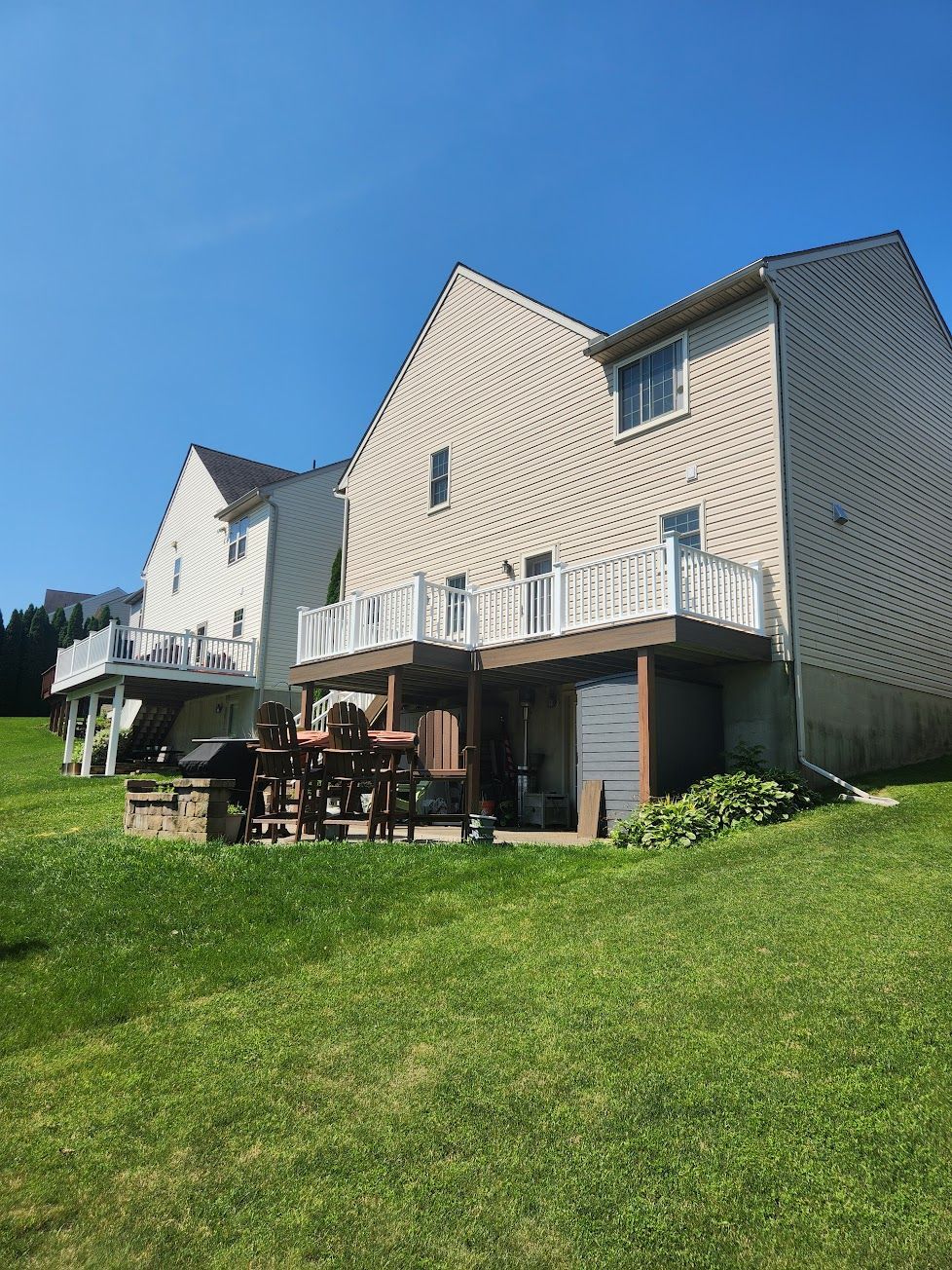 Two multi-story houses with beige siding and wooden decks, situated on a grassy hill under a clear blue sky.