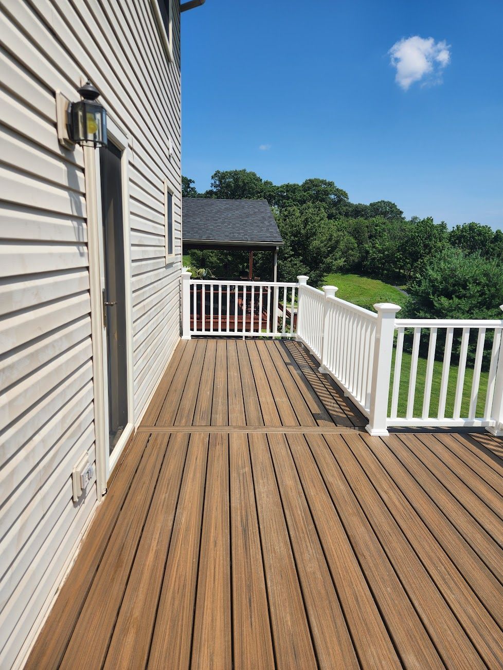 A brown deck with a white railing attached to the side of a light-colored house overlooking a tree-filled landscape.