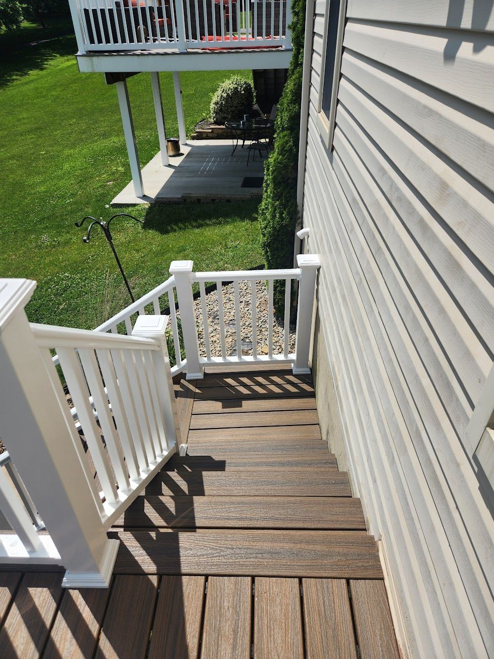 Wooden deck stairs with white railings lead down alongside light-colored vinyl siding to a grassy backyard patio.