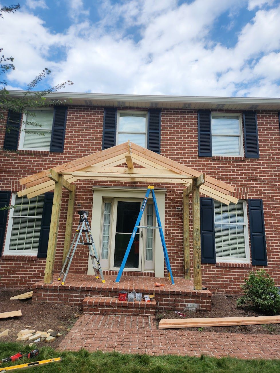 A wooden porch frame is being built over the front door of a two-story brick house with a blue ladder resting inside.