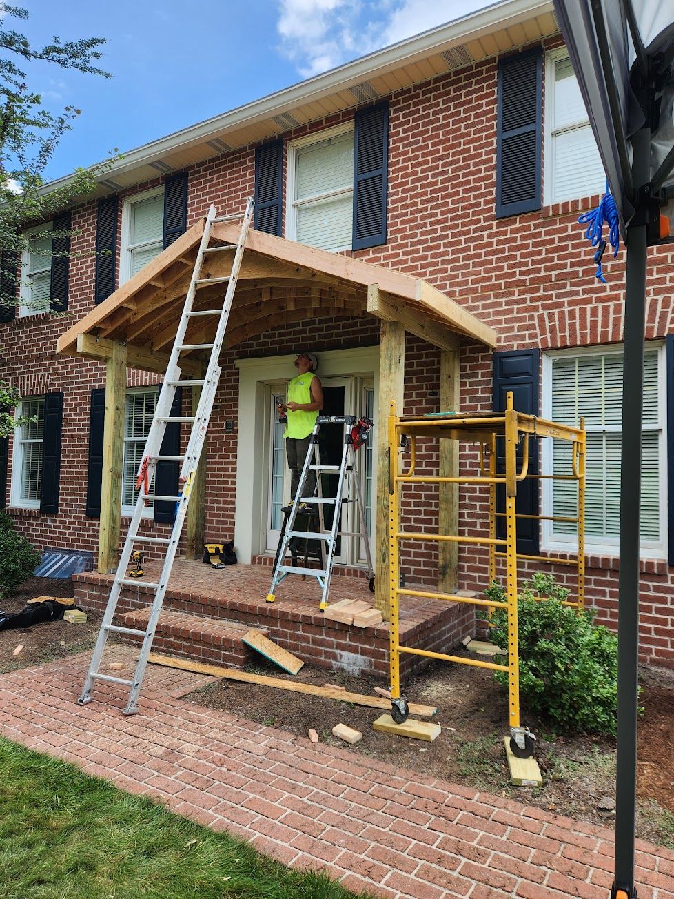 A person in a bright yellow shirt stands on the front porch of a brick house under a new wooden roof frame under construction.