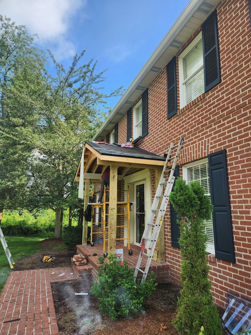 A partially constructed wooden porch roof attached to a red brick house, with a metal ladder leaning against the wall.